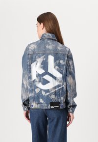 Denimjakke med en falmet, slitt look. Har en stor hvit logo på baksiden og tekst med "KARL LAGERFELD JEANS". Blå knapper, standard krage.