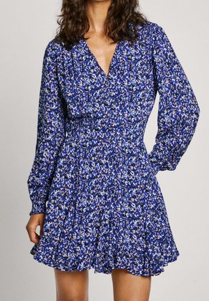 Vrouw die een blauwe bloemenjurk met lange mouwen, een V-hals en een uitlopende rok draagt, staand met één hand op haar heup.