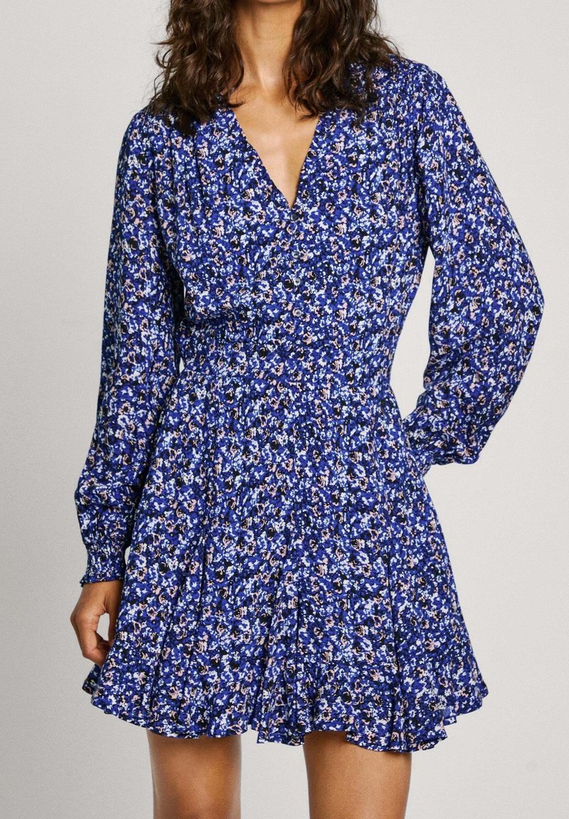 Femme portant une robe bleue fleurie à manches longues avec un décolleté en V et une jupe évasée, debout avec une main sur la hanche.