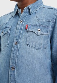 Jasnoniebieska koszula z denimu z kołnierzykiem na guziki, dwoma kieszeniami na klatce piersiowej, srebrnymi akcentami i małą czerwoną metką marki na lewej kieszeni.