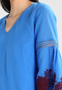 Blauwe linnen blouses met een V-hals, voorzien van geborduurde kastanjebruine bloemenpatronen op de mouwen en een kanten afwerking bij de manchetten.