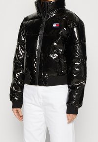 Osoba oblečená v lesklé černé péřové bundě s nášivkou Tommy Jeans a bílých kalhotách na jednotném pozadí.