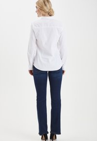 Camicia bianca con bottoni, maniche lunghe, pince sul retro e finitura pulita, abbinata a jeans svasati blu scuro e tacchi alti.