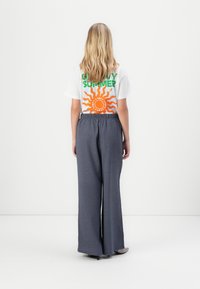 T-shirt blanc avec texte "SUMMER" en vert et orange accompagné d'un graphique de soleil, associé à un pantalon large ample gris foncé à motif et des chaussures à talons.