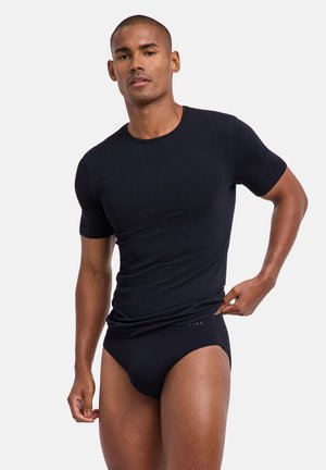 Mann trägt ein eng anliegendes schwarzes T-Shirt mit kurzen Ärmeln und passende schwarze Slips, richtet den Bund, steht vor einem schlichten hellen Hintergrund.