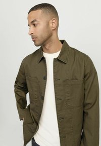 Giacca utility verde oliva in cotone, con tasche frontali, chiusura a bottoni e vestibilità rilassata, indossata da un modello con abbigliamento chiaro.
