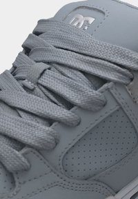 Primo piano dei lacci di una sneaker grigia annodati sopra il pannello laterale perforato con logo DC in rilievo sulla linguetta.