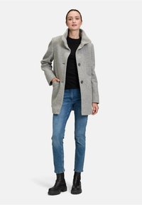 Cappotto grigio in misto lana con colletto a ribaltina, due tasche frontali e bottoni neri. Indossato sopra una maglietta nera e jeans blu con stivaletti neri.
