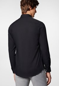 Czarna koszula z długim rękawem o zaokrąglonym dolnym brzegu, z zapięciem na guziki przy mankietach i gładką teksturą, widziana z tyłu.