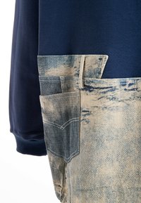 Sudadera azul marino con un bolsillo de denim claro texturizado. El bolsillo presenta una costura curva y costuras visibles, con un aspecto desgastado.