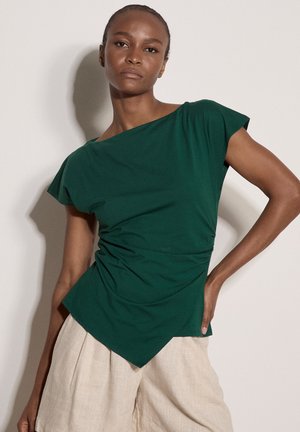 Femme aux cheveux courts portant un haut asymétrique à manches courtes vert foncé et un pantalon beige à jambes larges, debout avec une main sur la hanche.
