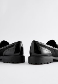 Schwarze Budapester Schuhe mit glänzendem Lacklederoberteil, verziert mit Brogue-Details und strukturierten Gummisohlen, die eine klassische Silhouette zeigen.