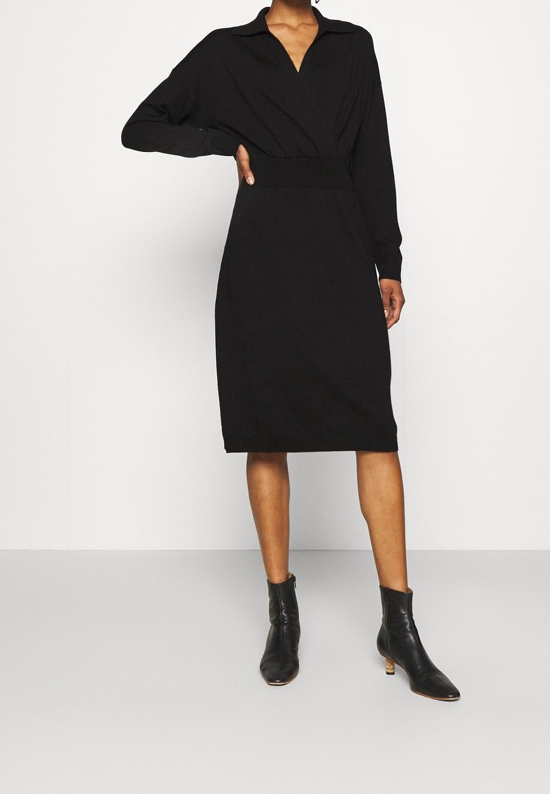 Robe noire à manches longues avec col, taille cintrée, et design au niveau du genou. Portez avec des bottines noires. Texture de tissu lisse.