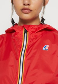 Cazadora roja con cierre delantero, capucha y logo de K-Way; presenta un diseño a rayas en el cierre en amarillo, negro y blanco.