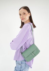 Grøn læder crossover taske med struktureret overflade, justerbar skulderrem og en frontflap. Båret sammen med en lys lilla top og blå jeans.