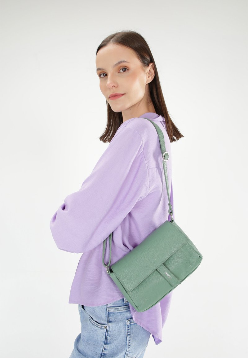 Zelená kožená crossbody taška s texturovaným povrchem, nastavitelného popruhu a přední klopou. Nosená s světlým fialovým topem a modrými džíny.