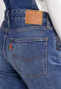 Denim overalls in faded blue, featuring a leather brand patch, stitched back pockets, and signature orange tab on the side.