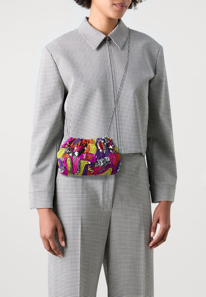 Personne portant une chemise et un pantalon gris à carreaux, portant un sac bandoulière coloré et brodé avec des sequins et des motifs abstraits.