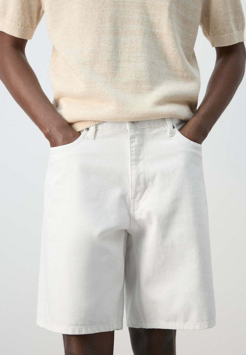 Personne portant un short blanc ample au genou et un haut en maille beige à manches courtes, les mains reposant dans les poches avant du short.