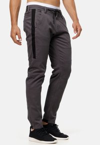 Gray cotton pants with black side stripes, tapered fit, drawstring waist, and zipper pockets. Worn with black sneakers and a white waistband.