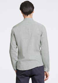 Camicia a maniche lunghe, in tessuto verde chiaro testurizzato, orlo arrotondato, polsini con bottone e colletto stretto, indossata con pantaloni scuri. Vista posteriore mostrata.