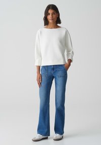 Pull en maille blanc avec un col bateau, associé à un jean bleu évasé comportant des poches avant et une coupe décontractée. Des baskets blanches viennent compléter le look.