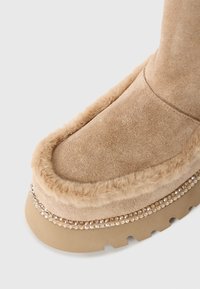 Beige suedestøvlett med myk pelskant og en rekke små glitrende rhinestener over den tykke gummisålen.