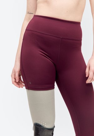 Personne portant un short bordeaux et une jambe prothétique grise avec une housse noire texturée, debout devant un fond blanc.