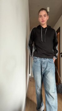 Sudadera negra con mangas grises, jeans azules holgados y una bolsa negra con lunares blancos. Ambiente interior con suelo de madera clara.