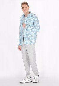 Lichtblauwe gemuste jas met een capuchon, voorzien van bloemmotieven, met ritssluitingen in de zakken, gecombineerd met grijze joggingbroeken en witte sneakers.