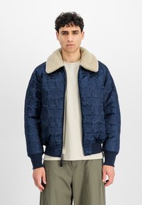 Marineblå quiltet bomberjakke med en beige faux shearling krave, ribbede manchetter og en frontlynlås. Båret over en lys skjorte.
