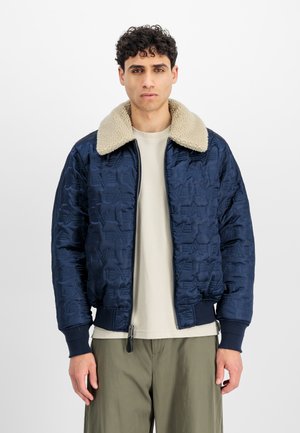 Marineblå quiltet bomberjakke med en beige faux shearling krave, ribbede manchetter og en frontlynlås. Båret over en lys skjorte.