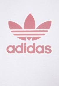 Biała tkanina z czerwonym, perforowanym logo Adidas, przedstawiającym trzy listki koniczyny nad nazwą marki w pogrubionej, zaokrąglonej czcionce.