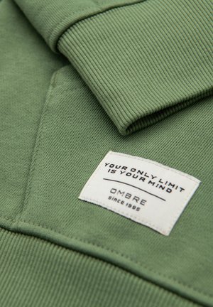 Manche de sweat-shirt vert et poche avec une étiquette en tissu blanc portant l'inscription « VOTRE SEULE LIMITE EST VOTRE ESPRIT » et « OMBRE depuis 1989 ».