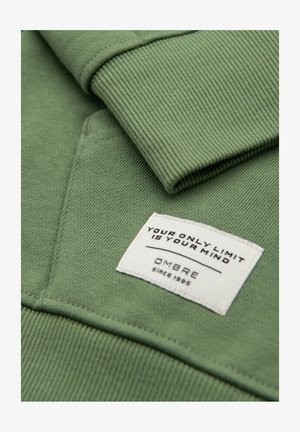 Grüner Sweatshirtärmel und Tasche mit einem weißen Stoffetikett mit der Aufschrift "DEINE EINZIGE GRENZE IST DEIN GEIST" und "OMBRE seit 1989."