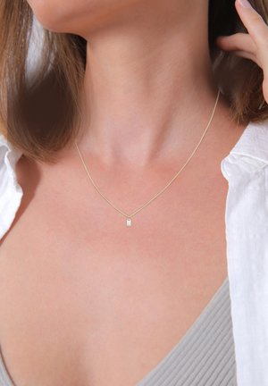 Goldenes Halskette mit einer zarten Kette und einem kleinen rechteckigen Anhänger, der einen klaren Edelstein trägt. Getragen über einem hellgrauen Oberteil.