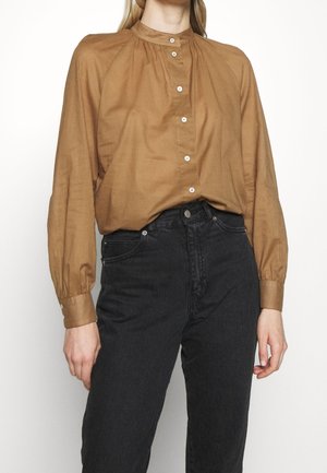 Woman wearing a brown long-sleeve button-up blouse with puffed sleeves tucked into high-waisted black jeans.