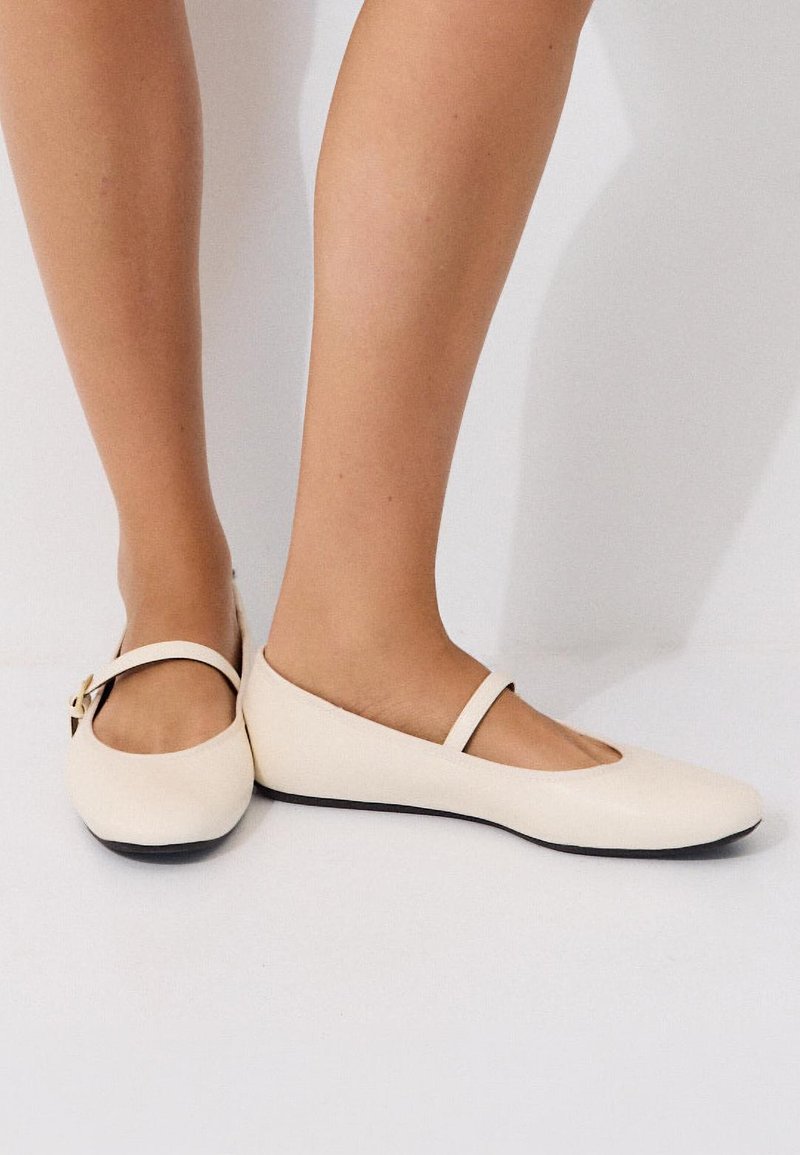 Pieds portant des ballerines Mary Jane couleur crème avec une fine bride et des semelles noires sur fond blanc.