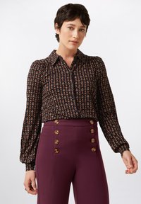 Schwarze Bluse mit floralem Muster, langen Ballonärmeln und einer Knopfleiste, kombiniert mit hoch taillierten, kastanienfarbenen Hosen mit goldenen, runden Knöpfen.