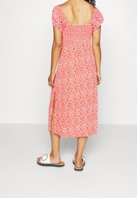 Robe d'été rouge à fleurs avec manches courtes bouffantes, corsage froncé et fentes latérales. Portée avec des sandales blanches comportant un accent noir.