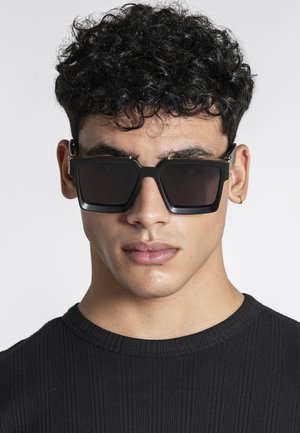 Mladić s kovrčavom crnom kosom koji nosi velike crne kvadratne sunčane naočale i malu zlatnu obruču u uhu, obučen u crnu rebrastu majicu.