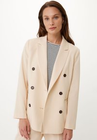 Blazer beige doppiopetto realizzato in tessuto liscio, con grandi bottoni scuri e un collo strutturato, indossato sopra una maglietta a righe.