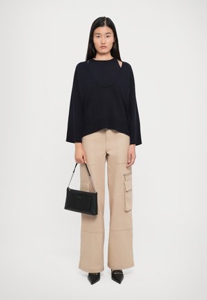 Woman stands against white background wearing black wide-sleeve top, beige cargo pants, black pointed shoes, holding black handbag in one hand.