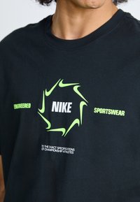 Pessoa a usar uma t-shirt preta com logótipos da Nike em verde néon, formando um círculo em torno do texto "NIKE" e a legenda "Engineered Sportswear".