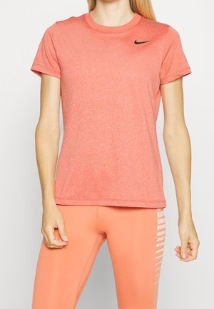 Femme portant un t-shirt Nike corail à manches courtes et des leggings corail avec un texte blanc sur le côté, debout les bras le long du corps sur un fond uni.