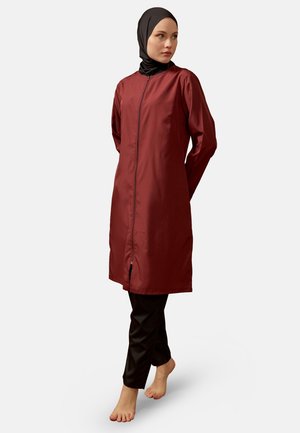 Woman wearing a black hijab, long maroon zip-up tunic, and black pants, standing barefoot on a white background, looking to the side.