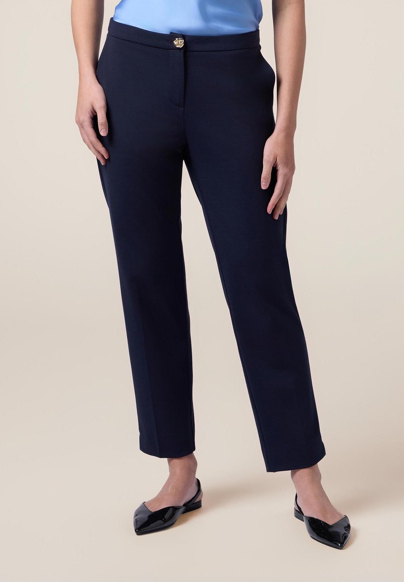 Pantalons ajustés bleu marine à coupe droite, avec une fermeture à bouton sur le devant et des poches. Portés avec des ballerines noires à bouts pointus.