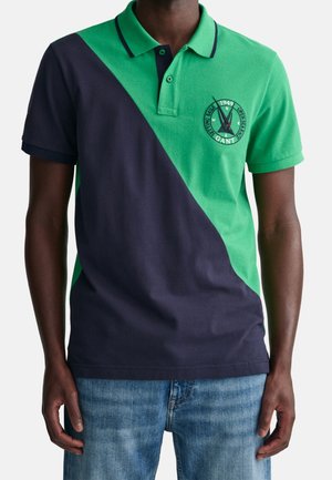 Man wearing a short-sleeve polo shirt with diagonal green and navy color blocks and a circular embroidered logo on the chest.