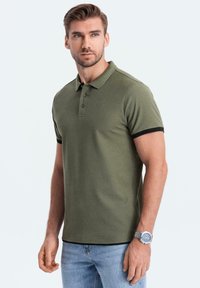 Olive zelené polo tričko s černým žebrovaným límcem a manžetami, s třemi knoflíky vpředu, v kombinaci se světle modrými denimovými džíny.
