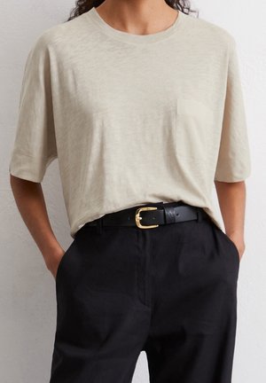 Person iført en beige kortærmet skjorte med brystlomme, der er stukket ind i sorte bukser med et sort bælte med en guldbetrukket spænde.